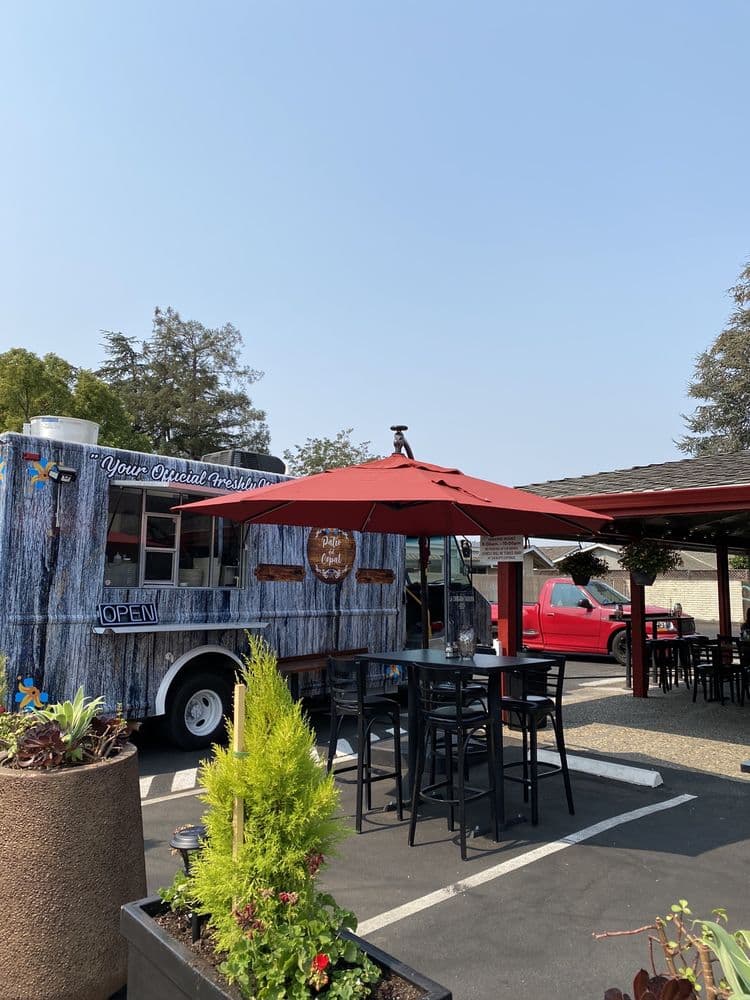The blue-wrapped kitchen truck parked beside the patio, with a red umbrella and bar tables